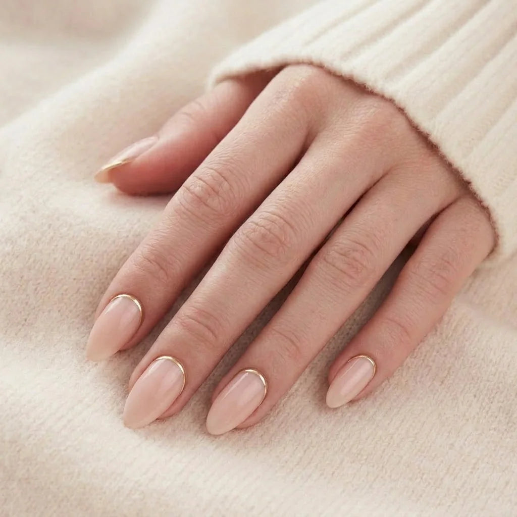 En hand med nudefärgade press-on naglar i mandelform med tunn guldkant vid nagelbandet, mot en stickad tröja