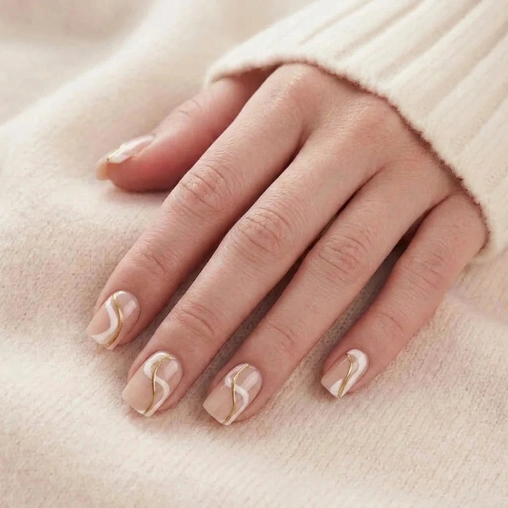 Korta, beige lösnaglar med vit och guldig vågdesign på en modellhand mot stickad tröja.