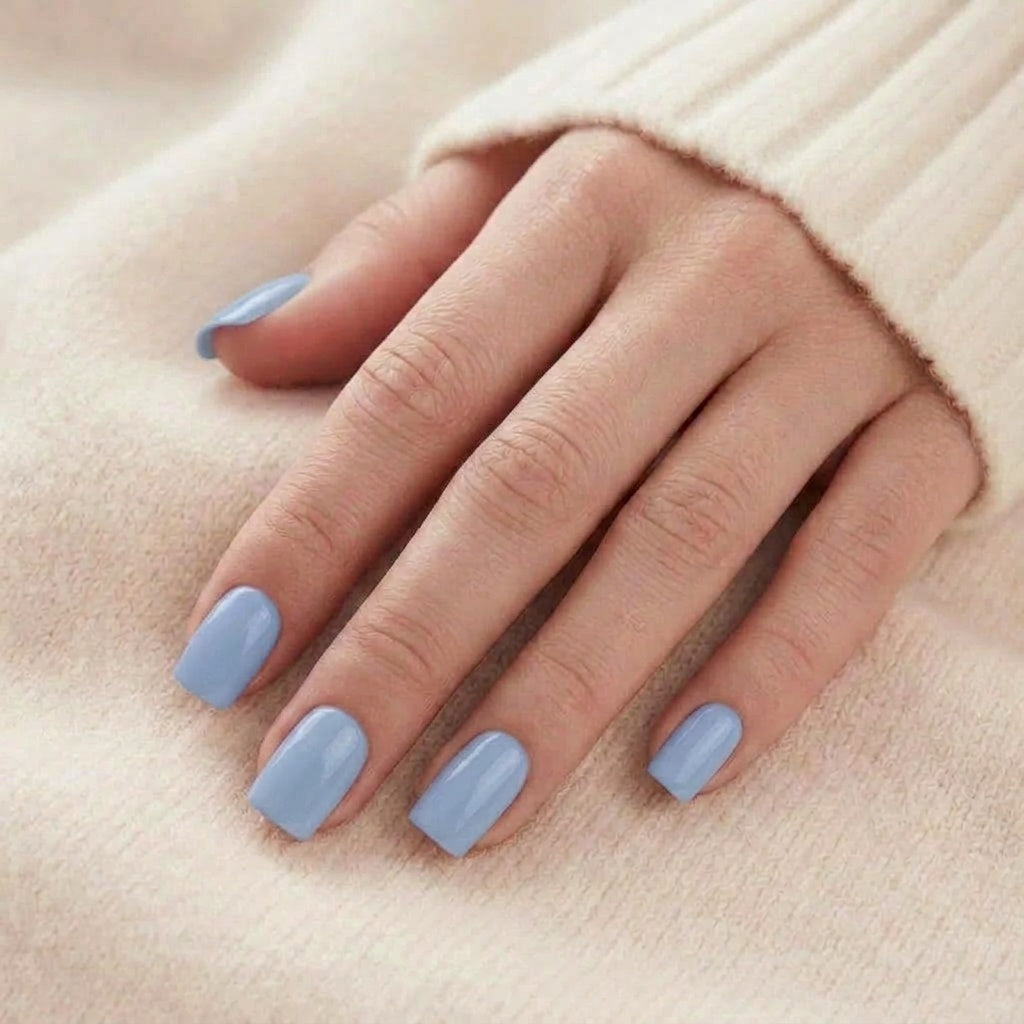 Baby Blue press-on naglar i ljusblå, kort fyrkantig form med glansig yta, hand vilar på beige stickat tyg